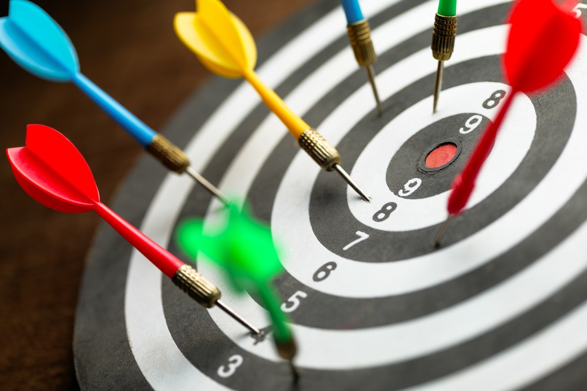 A close-up of seven darts on a dartboard, grouped around the center but all missing the bullseye.